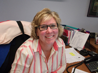 woman with blonde hair and glasses, smiling at her desk